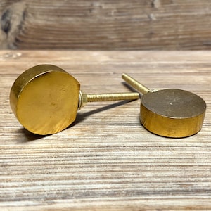 Może przedstawiać: Dwa złote gałki do szafek o płaskim, okrągłym kształcie. Każda gałka ma gwintowaną śrubę do mocowania. Gałki wykonane są z litego metalu i są prezentowane na drewnianej powierzchni.