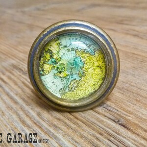 May include: A round cabinet knob with a vintage map design. The knob features a brass-colored metal base and a clear glass dome over the map. The map shows the Arctic region in shades of green, yellow, and blue. The knob is on a wooden surface.