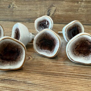 May include: A collection of decorative cabinet knobs. Each knob features a white ceramic base with a brown and purple center, framed by a gold-colored edge. The knobs are arranged on a wooden surface.
