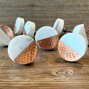 May include: A collection of decorative cabinet knobs. Each knob features a half-circle of white marble and a half-circle of hammered copper. The knobs are round with a metal screw for installation. The knobs are on a wooden surface.