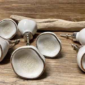 Könnte beinhalten: Eine Sammlung weißer Keramik-Schrankknöpfe mit Craquelé-Effekt und silberfarbener Hardware. Die Knöpfe haben eine einzigartige, organische Form mit goldfarbenem Rand. Diese dekorativen Knöpfe eignen sich ideal zum Aufwerten von Möbeln.
