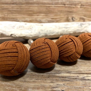Pode incluir: Quatro esferas decorativas de couro castanho dispostas em fila. Cada esfera é feita de tiras de couro trançadas, criando uma forma texturizada semelhante a uma bola. As esferas estão sobre um fundo de madeira rústica. Cada esfera mede cerca de 5 cm de diâmetro.