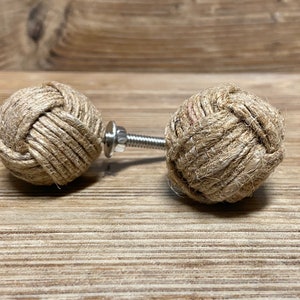 以下が含まれることがあります： 2つの装飾的なキャビネットノブ。各ノブは、天然色のロープをきつく織った球体です。銀色のネジとナットがロープの球体をつないでいます。ノブは、明るい茶色の木製の表面に表示されています。