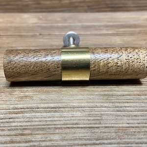 Pode incluir: Um puxador de armário de madeira com design cilíndrico. O puxador apresenta um corpo de madeira clara e texturizada com uma faixa de latão ao redor do centro. Um parafuso prateado é fixado na parte traseira para instalação.