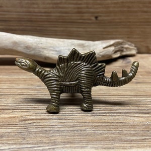 Op de afbeelding: Een bronzen dinosaurusbeeldje met een getextureerd oppervlak. De dinosaurus staat op een houten oppervlak.