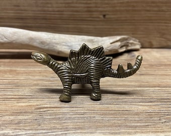May include: A bronze-colored dinosaur figurine with a textured surface. The dinosaur is standing on a wooden surface.