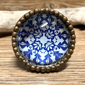 May include: A round cabinet knob featuring a blue and white floral pattern under a clear glass dome. The knob is encased in a textured, antique bronze-coloured metal frame with a beaded edge, providing a decorative accent for furniture hardware.