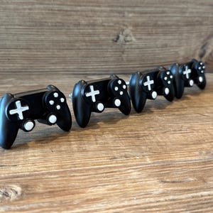 May include: A series of black cabinet knobs in the shape of game controllers, featuring white directional pads and buttons. The knobs are displayed on a wooden surface, providing a gaming-themed decorative touch.