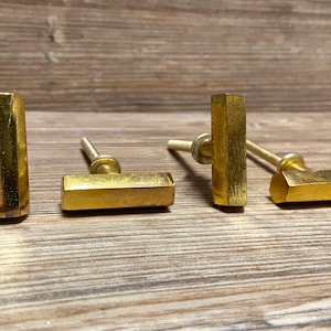 Op de afbeelding: Vier goudkleurige kastknoppen met een rechthoekig staafontwerp en een zeshoekige vorm. De knoppen zijn gemaakt van metaal en hebben een messing afwerking. De hardware wordt getoond op een houten oppervlak.