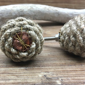 Peut inclure: Deux boutons de meuble décoratifs. Chaque bouton est fait de corde naturelle étroitement enroulée, avec un accent circulaire en cuivre et laiton au centre. Les boutons sont fixés à une vis métallique. Les boutons sont sur une surface en bois.