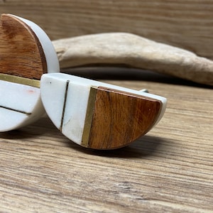 May include: Two half-circle cabinet knobs. Each knob features a combination of materials: white marble, dark wood, and brass accents. The knobs are designed with a modern aesthetic, suitable for various furniture styles.