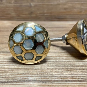 May include: Two decorative cabinet knobs. Each knob has a gold-coloured metal base with a hexagonal pattern filled with iridescent, pearlescent white and light blue accents. The knobs are on a wooden surface.