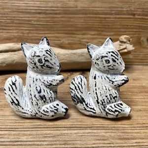 May include: Two white ceramic squirrel figurines with black and white striped patterns. The squirrels are sitting upright with their tails curled behind them.