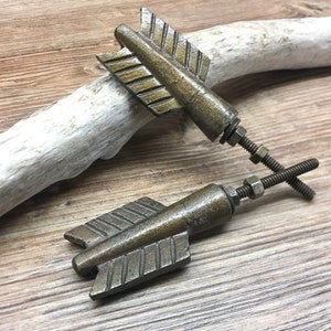 May include: Two antique brass-toned metal drawer knobs shaped like arrow fletching. Each knob features a textured, triangular fin design with a threaded metal bolt, a washer, and a nut attached to the back for secure installation on furniture.