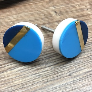 May include: Two circular cabinet knobs with a contemporary design. Each knob has a white base, a section of navy blue, and a section of light blue, divided by a gold-coloured stripe. The knobs are mounted on silver screws.