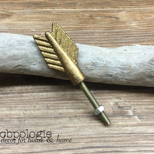 May include: A gold-toned metal cabinet knob shaped like an arrow fletching. The knob features a textured, weathered finish and includes a threaded metal bolt with a single hexagonal nut attached to the base for secure installation on furniture.