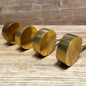 May include: Four brass cabinet knobs with a rounded, flat-faced design. The knobs are arranged in a row, showcasing their warm, golden color and simple, modern aesthetic. Each knob has a screw for easy installation.