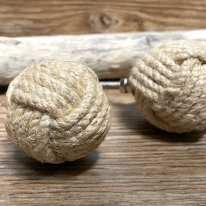 Peut inclure: Deux boutons de meuble sphériques décoratifs en corde naturelle étroitement enroulée. Chaque bouton présente un motif de nœud et est fixé à une vis et un écrou métalliques. L'ensemble est présenté sur un fond en bois, évoquant un thème nautique ou rustique.