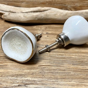 Könnte beinhalten: Zwei weiße Keramik-Schrankknöpfe mit Craquelé-Design und goldfarbenem Rand. Die Knöpfe haben eine abgerundete Form und sind an silberfarbener Metallbeschlägen befestigt. Sie werden auf einer Holzoberfläche präsentiert.