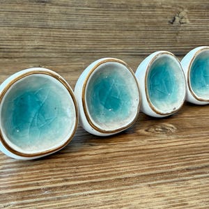 May include: Four white ceramic cabinet knobs with a turquoise interior and gold trim. The knobs have a smooth, rounded shape and are arranged in a row on a wooden surface. The turquoise colour has a crackle effect.