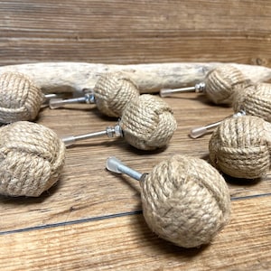 Peut inclure: Ensemble de dix boutons de meubles décoratifs. Chaque bouton est composé d'une sphère enveloppée de corde naturelle, fixée à une vis métallique avec un embout en plastique transparent. Présentés sur une surface en bois.