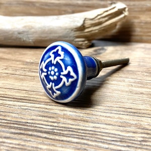 Può includere: Pomello per armadio in ceramica blu e bianca con un motivo floreale. Il pomello ha una forma rotonda con un motivo floreale bianco su sfondo blu. L'hardware è di colore argento.