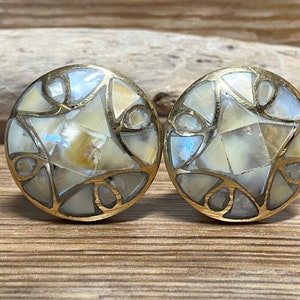 May include: A pair of round cabinet knobs with a mosaic design. Each knob features iridescent, cream-coloured shell pieces set within a gold-toned metal frame. The knobs are displayed on a wooden surface, highlighting their intricate details and craftsmanship.