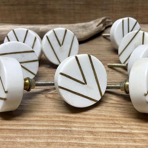 Könnte beinhalten: Eine Sammlung weißer Marmor-Schrankknöpfe mit messingfarbenen geometrischen Designs. Jeder Knopf hat eine runde Oberfläche mit einer messingfarbenen Schraube, vor einem Holzgrund.