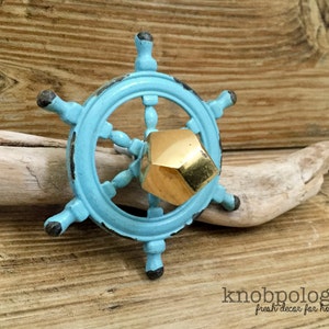 May include: A nautical-themed cabinet knob in the shape of a ship's wheel, painted in a distressed turquoise finish. The center features a gold-colored, geometric knob. The knob is set against a wooden background.