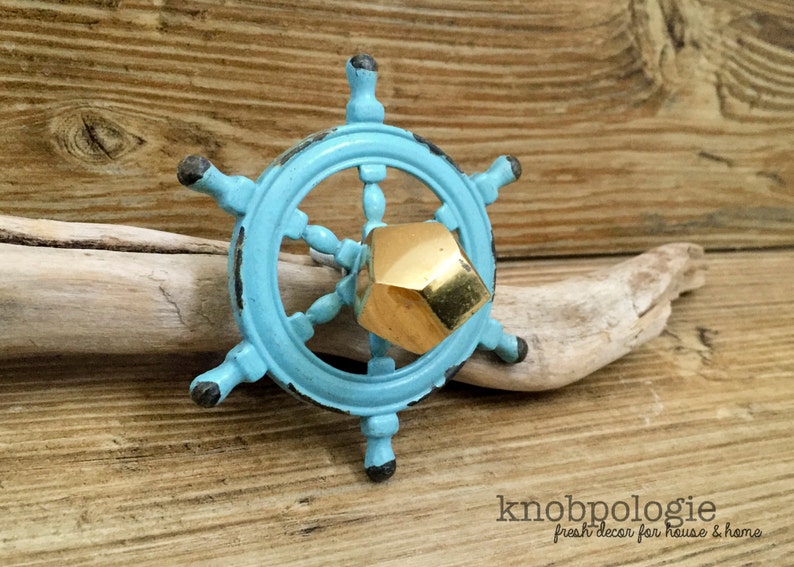 May include: A nautical-themed cabinet knob in the shape of a ship's wheel, painted in a distressed turquoise finish. The center features a gold-colored, geometric knob. The knob is set against a wooden background.