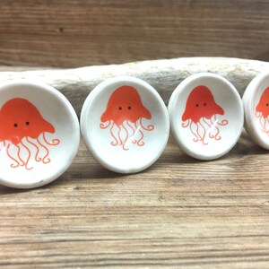 Puede incluir: Cuatro pequeños cuencos de cerámica blanca con un diseño de medusa naranja. Las medusas tienen una campana redondeada y tentáculos. Los cuencos están dispuestos sobre una superficie de madera, sugiriendo un tema náutico.
