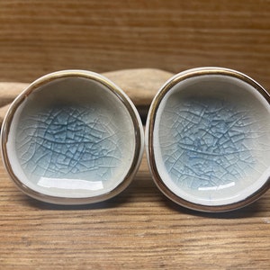 May include: Two small, ceramic bowls with a crackle glaze in shades of blue and white. The rims are trimmed with a gold-colored band. The bowls are displayed on a wooden surface.