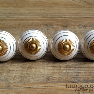 May include: Four white cabinet knobs with gold accents. Each knob has a gold center and gold concentric circles. The knobs are arranged in a row on a wooden surface. The knobs are a decorative element for furniture.