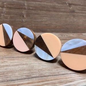 Pode incluir: Quatro puxadores redondos para armários com um design geométrico. Cada puxador apresenta uma secção de madeira escura, uma secção de material claro e uma secção de cor sólida, como pêssego ou branco. Os puxadores estão dispostos numa superfície de madeira.