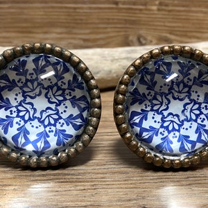 May include: Two circular cabinet knobs featuring a blue and white floral pattern under a clear glass dome. Each knob is encased in a textured, antique bronze-coloured metal frame with a beaded edge, providing a distinct contrast to the blue design.