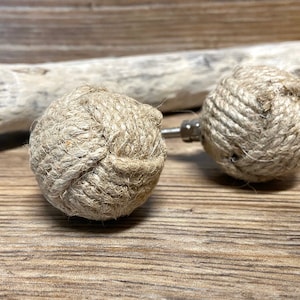 May include: Two decorative cabinet knobs. Each knob features a sphere wrapped in natural-colored rope, attached to a silver-toned metal base. The knobs are set against a wooden background, suggesting a nautical or rustic theme.