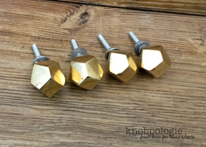 May include: Four gold-colored, faceted cabinet knobs with silver screws are arranged on a wooden surface. The knobs have a geometric, gem-like design. The knobs are for home decor.