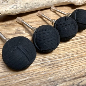 Può includere: Quattro pomelli neri per armadi con un design rotondo e strutturato. Ogni pomello ha uno stelo in metallo argentato e una punta trasparente e arrotondata. I pomelli sono disposti su una superficie di legno.