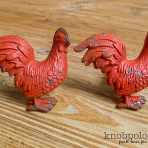 Puede incluir: Dos tiradores de armario rojos con forma de gallo y pintura desgastada. Los tiradores son de metal y tienen un aspecto rústico y vintage. Los tiradores están diseñados para su uso en muebles y añaden un toque decorativo.