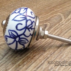 Può includere: Pomello per mobili in ceramica bianca e blu a motivi floreali, con base e vite in metallo argentato. Il pomello ha una forma arrotondata ed è progettato per mobili. Il testo "knobpologie" è visibile.