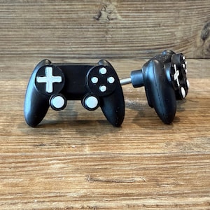 May include: Two black controller-shaped cabinet knobs with white buttons and a white cross. The knobs are mounted on a silver screw. The knobs are on a wooden surface.