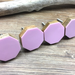 May include: Four decorative cabinet knobs with a light purple, hexagonal face and a wooden base. The knobs are set against a light brown wooden surface, with a piece of driftwood in the background.