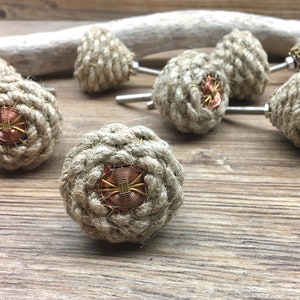 May include: A set of rustic cabinet knobs. Each knob is made from tightly wound natural rope, with a copper-coloured centre and a decorative wire design. The knobs are mounted on silver-toned screws, ideal for adding a nautical touch to furniture.