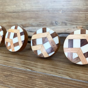 Op de afbeelding: Vier ronde houten knoppen met een geometrisch patroon van wit, bruin en lichtbruin hout. De knoppen zijn in een rij op een houten oppervlak gerangschikt.