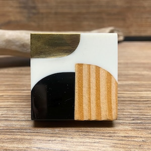 May include: Square tile with a geometric design featuring a black semi-circle, a gold semi-circle, and a striped brown semi-circle.