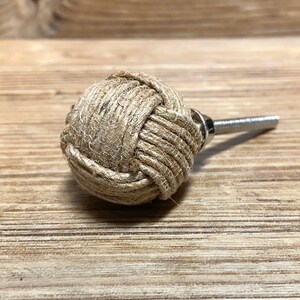 May include: A decorative cabinet knob with a natural jute rope design. The round knob is tightly woven and attached to a silver-colored metal screw. The knob is approximately 1.5 inches in diameter, perfect for adding a rustic touch to furniture.