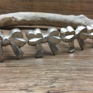 May include: Six silver-colored bow-shaped cabinet knobs arranged in a row. The knobs are made of metal and have a detailed, textured design. They are set against a wooden background with a light-colored, elongated object in the background.