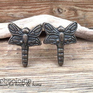 May include: Two antique bronze dragonfly drawer pulls with a detailed design. The pulls are mounted on a wooden surface.