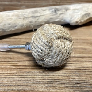 Peut inclure: Bouton de meuble décoratif avec un design sphérique en corde naturelle étroitement enroulée. Le bouton est fixé à un boulon métallique avec un écrou et un embout en plastique transparent. L'arrière-plan comprend un morceau de bois flotté.