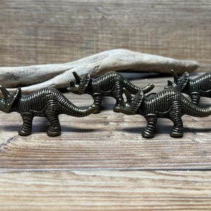 May include: Four bronze-coloured dinosaur-shaped cabinet knobs. The dinosaurs are in a line, facing the same direction. The dinosaurs are detailed with textured skin and horns.